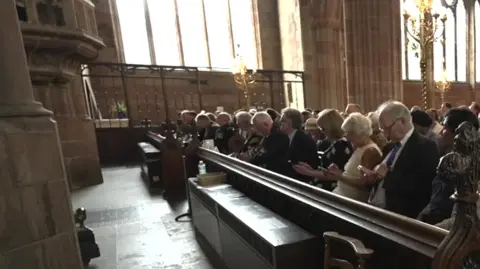 The congregation are seated in the middle and on the right of the photo, facing towards the left on the image. Church windows are in the background, with bright light coming through them.