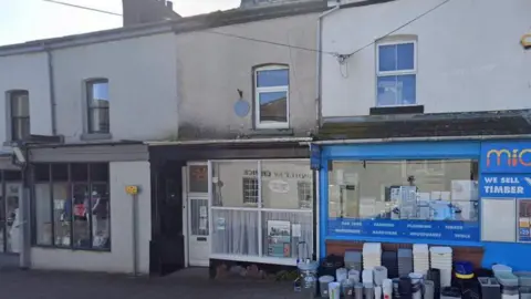 A row of terraced shops, with Norman Nicholson's former home in the centre, which has glass windows with a netting curtain. 