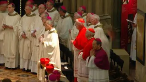 Lancaster Cathedral The future pope is in the foreground of a line of priests in their vestments in front of two two cardinals in red skull caps. The sun is shining on him.