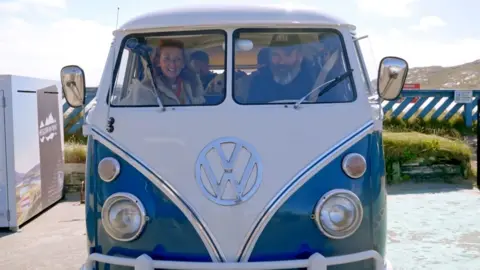 Solus Productions/BBC Jules and Gregg are seated in front in a blue and white vintage Volkswagen campervan on a sunny day. Their two sons, both in their early twenties, are positioned in the rear of the vehicle.