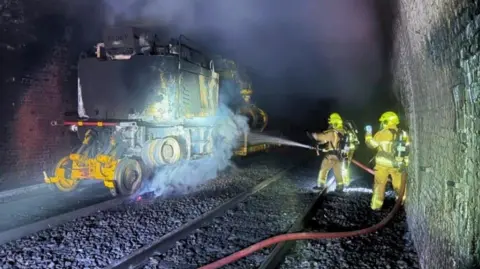 A maintenance vehicle in Standedge Tunnel being hosed down by firefighters wearing protective clothing 