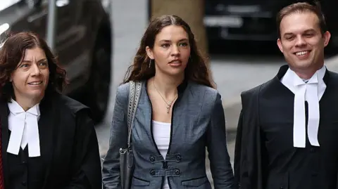 Getty Images A young woman with long brown hair, wearing a white top and grey jacket, flanked by two barristers in black and white legal gowns, walking down a street