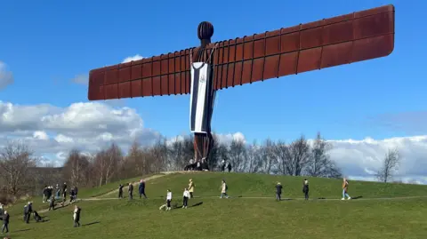 El ángel del norte cuelga de su cuerpo con una camisa de Newcastle United en blanco y negro. La gente se ve debajo de las montañas.