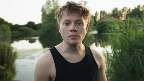 Create Studios A man in a black sleeveless top stands, with a serious expression. He is stood in front of a lake, surrounded by green trees and foliage. The water reflects the natural scenery. 
