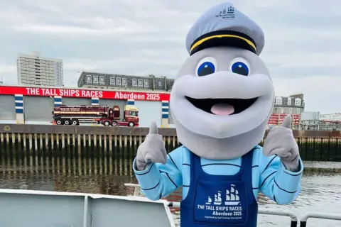 Dorry the dolphin mascot on a boat, giving thumb up, with sign saying 'Tall Ships Races Aberdeen 2025' in the background.