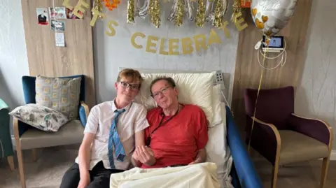 Supplied Beckett holds his dad's hand in a care ward with celebrate balloons behind 