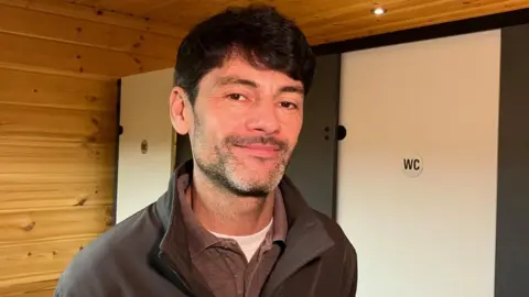 A man smiles at the camera, standing in a room with wood-panelled walls and cubicles with "WC" written on the doors.