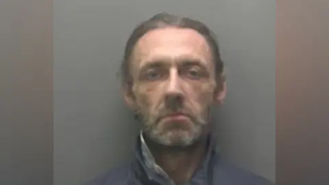 A police mugshot of James Conroy. He has pulled-back brown hair and a greying beard. He is wearing a dark grey anorak done up to his neck. 