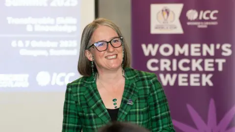 Sarah Gomersall stood in front of a screen and a purple screen. The purple sign has the words 'Women's Cricket Week'. She is wearing a black and green checked blazer with a black top underneath and a green necklace. 