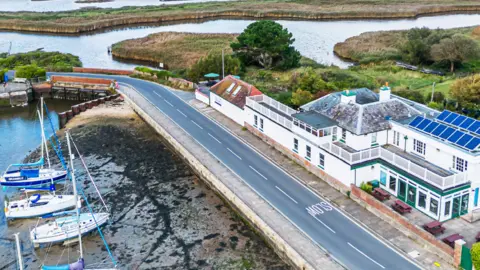 Aerial view of Haven House, which is across a road from yachts moored at a harbour. In the background are channels of water on either side of a thin strip of grassed land.