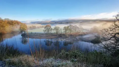 Robin Ree Mist and fog hangs over a river in front of a pebbled plateaued bank and green fields. Hills can be seen in the background alongside trees. The leaves on the trees topped with frost.