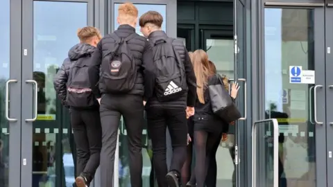 A group of three school boys dressed in black and carrying school bags walking through a school door