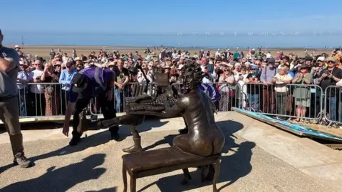 A bronze statue of Terry Jones as a naked organists in front of a crowd of people taking photos with the sea behind them