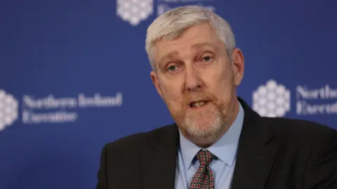 PA Media A close-up of John O'Dowd. He is an older man with short grey hair, a black blazer, a blue shirt and a patterned tie. He is standing in front of a blue NI Executive board and is speaking.