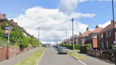 A hilly street with semi-detached houses in brick on either side and a number of parked cars
