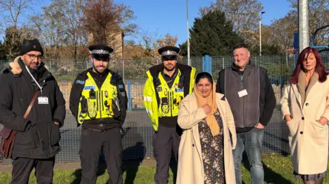 Cambridgeshire Constabulary Two police officers wearing high visibility coats and vests are stood in a park with two men and two women. 