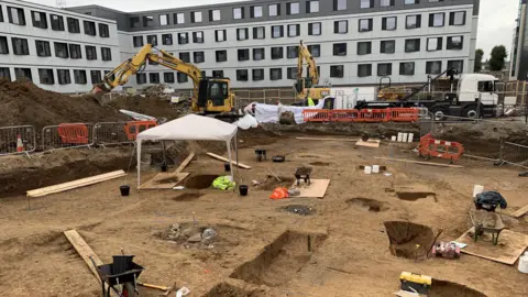 A square plot of land with piles of dirt and excavators at the back. Most of the plot looks meticulously dug out, with several archaeological trenches and various tools like wheelbarrows and spades dotted about.