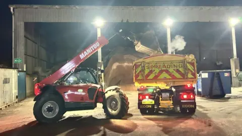 Dorset Council A vehicle with a crane deposits grit into the back of a lorry. A pile of sand is visible in the warehouse behind.