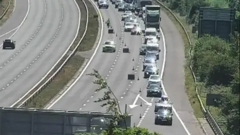 National Highways Motorway camera image of cars being sent off the motorway. There are long queues as cars and lorries make their way off the motorway with police and cones in view.