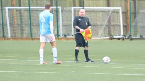 Warren Muggleton/BBC Stuart Nixon is in a black referee kit, holding a checked yellow and orange linesman's flag. He is on a green all-weather football pitch, talking to a man in a sky blue and white kit.