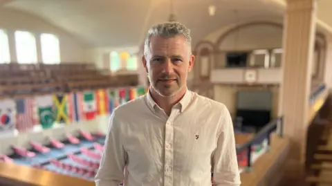 Mark Ansell/BBC Ben Gibbins with salt and pepper hair sporting a pink shirt and a three-day stubble. In the background are several flags mounted to the wall, including Jamaica's flag.