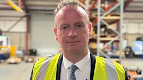 Stephen Kelly, a man with short, curly fair hair, looks at the camera in a factory or warehouse setting. He is wearing a yellow high-viz vest over a dark suit, white shirt and blue tie.