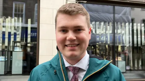 A man stands outdoors on a pedestrian street in front of a shopfront with large glass windows and vertical decorative panels. He's wearing a teal waterproof jacket with yellow trim over a white shirt and patterned tie.