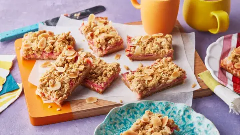A chopping board with an assortment of oat crumble bars sitting on top of it, next to some cups and bowls.