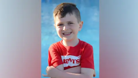 Archie York wears a red T-shirt saying Levis. He has dark hair, a big smile and is standing next to a swimming pool.