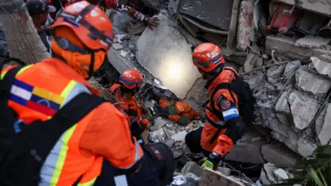 Reuters Three people in orange jackets with high-vis stripes and orange helmets can be seen from above as they descend into the rubble of a building. They are wearing head torches, which shine a light below.