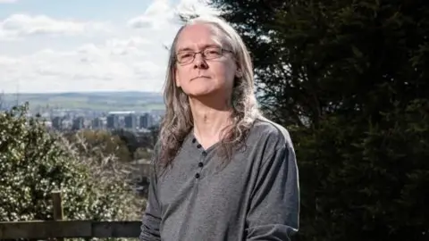 Martin Atkinson, a middle-aged man with long grey hair and glasses, stands looking serious on a grassed area. A cityscape is in the background. 