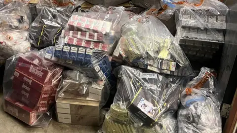 Several clear bags with seized cigarette packets and boxes in a pile on the floor.