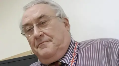 Supplied Leslie Irwin looking at the camera. He is an older man with grey hair and rimless glasses. He is wearing a purple and white striped shirt, a tie and a lanyard with a pattern of the Union Jack on. 