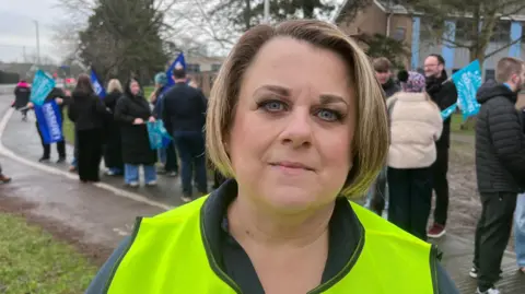 A woman in her 40s with a short dirty blonde bob. She is wearing makeup, a navy fleece, and a green hi-vis tabard as she stands in front of a picket line of teachers and staff outside a school. The teachers are wrapped up warm with coats and hats in the cold weather, and are holding up blue union flags.