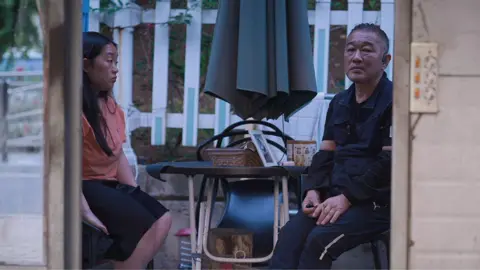 A man and woman sit at a small outdoor table beneath a closed umbrella on a patio with a white fence.