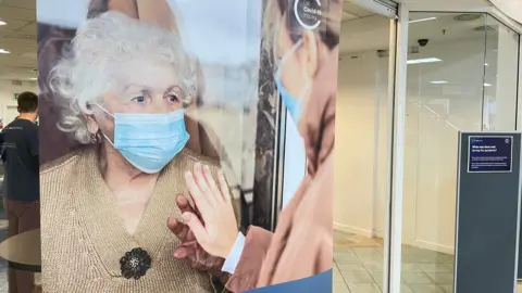 A large poster depicting an older woman touching a younger woman through glass while both a wearing face masks hangs in a window. The Covid inquiry branding is visible on a panel in the background. 