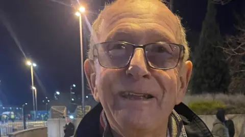 Plymouth Argyle supporter Jim smiles as he leaves the Etihad Stadium after watching the team lose 3-1 to Manchester City in the FA Cup fifth round. It is dark and several street lights are on in the background.