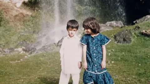 Khushal Yousafzai Khushal, wearing white outfit and Malala wearing a blue dress - as kids. They are standing in front of a waterfall, on green grass.