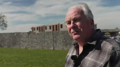 A man with white hair, swept back, stands on a green patch of land in front of a building site fenced off with high wooden walls. A large shell of a building can be seen in the background.