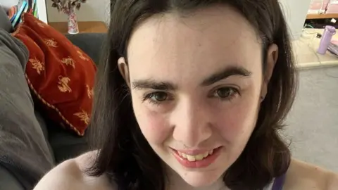 Dravet Syndrome UK Lucy looking at the camera and smiling in a selfie. There is a grey sofa behind her with a cushion on it.