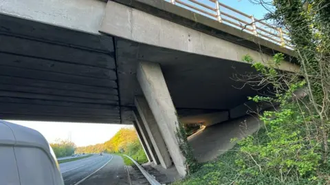 National Highways A view of underneath a concrete motorway bridge next to a three lane motorway carriageway.