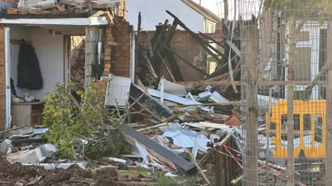 A house which has been burnt down to the ground with rubble surrounded by what is left of the house. The doorway to the house is still standing, and a coat rack and a chest of drawers can be seen in the entry way.