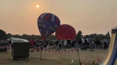 Neil Sackley Northampton balloon festival with hundreds of milling milling around two deflating hot-air balloons in a field at dusk.