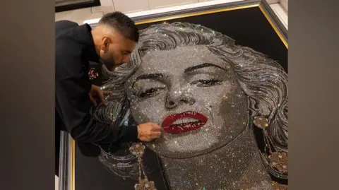 San B studios A portrait of Marilyn Monroe placed on the floor with an artist, who is wearing all black, hovering over it making final touches to her lips.
In the artwork Marilyn Monroe is wearing a gold top 