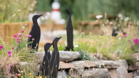 JANE PRESCOTT Small wooden silhouettes of birds are placed in the garden next to large, flat stone slates and long grass and flowers.