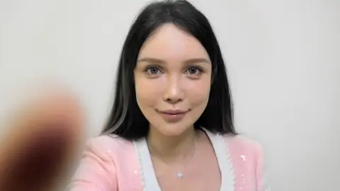 A young woman with a surgically-enhanced face, wearing a pink jumper, stares at the camera. White lines have been imposed over her face by a cosmetic surgery app called SoYoung 