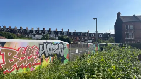 Graffiti on hoardings around the old Clifton Hill leisure centre site with overgrown weeds in the foreground and the backs of terraced houses in the background.