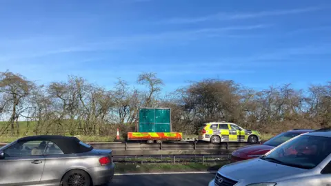 A police vehicle and emergency sign. Closer to the camera are several cars travelling on the nearside carriageway. 