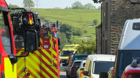 Fire engines and ambulances are at the scene in Ribchester. They are parked alongside other vehicles with a stone house to the right and green fields in the background on a sunny day. 