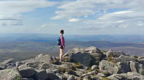 Ciaran McAteer A woman wearing shorts looking into the hilly distance from a rocky hilltop.